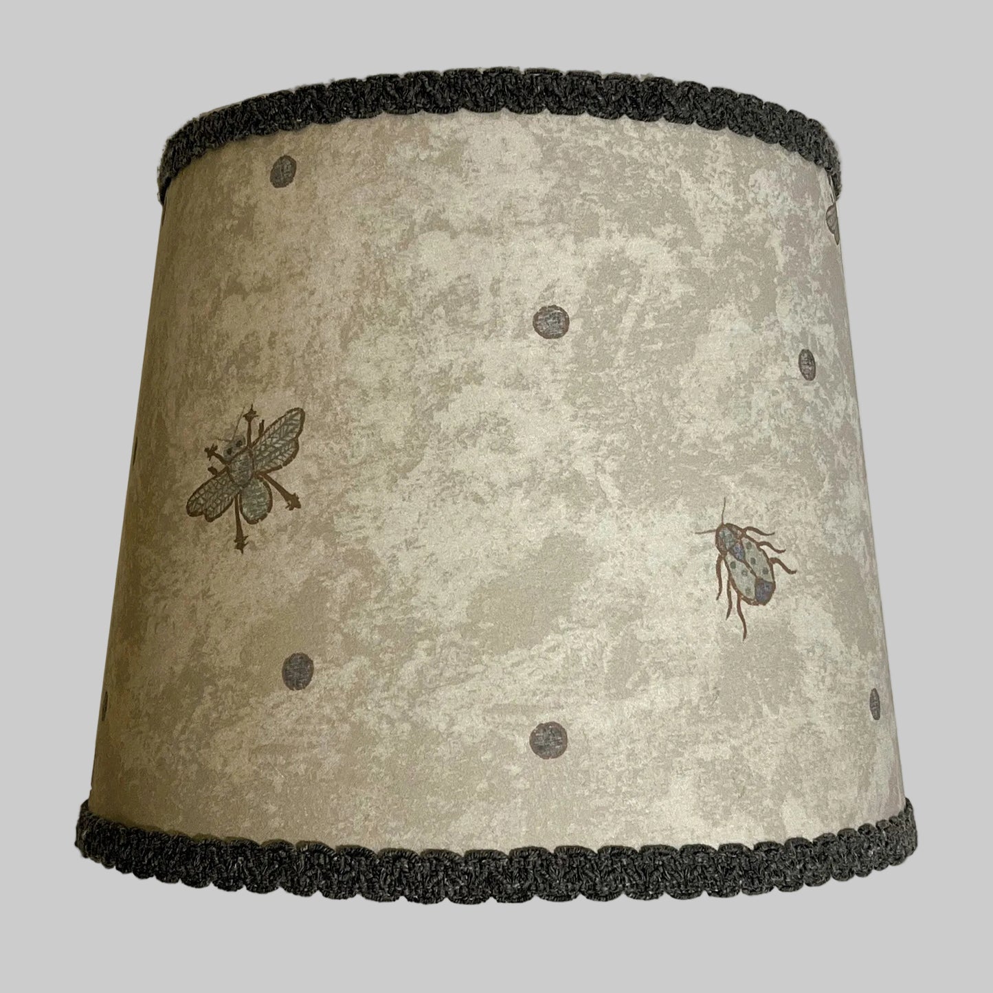 A lampshade with a beetle and spot pattern in an ocean color on a printed parchment background, with a dark grey gimp flat trim.